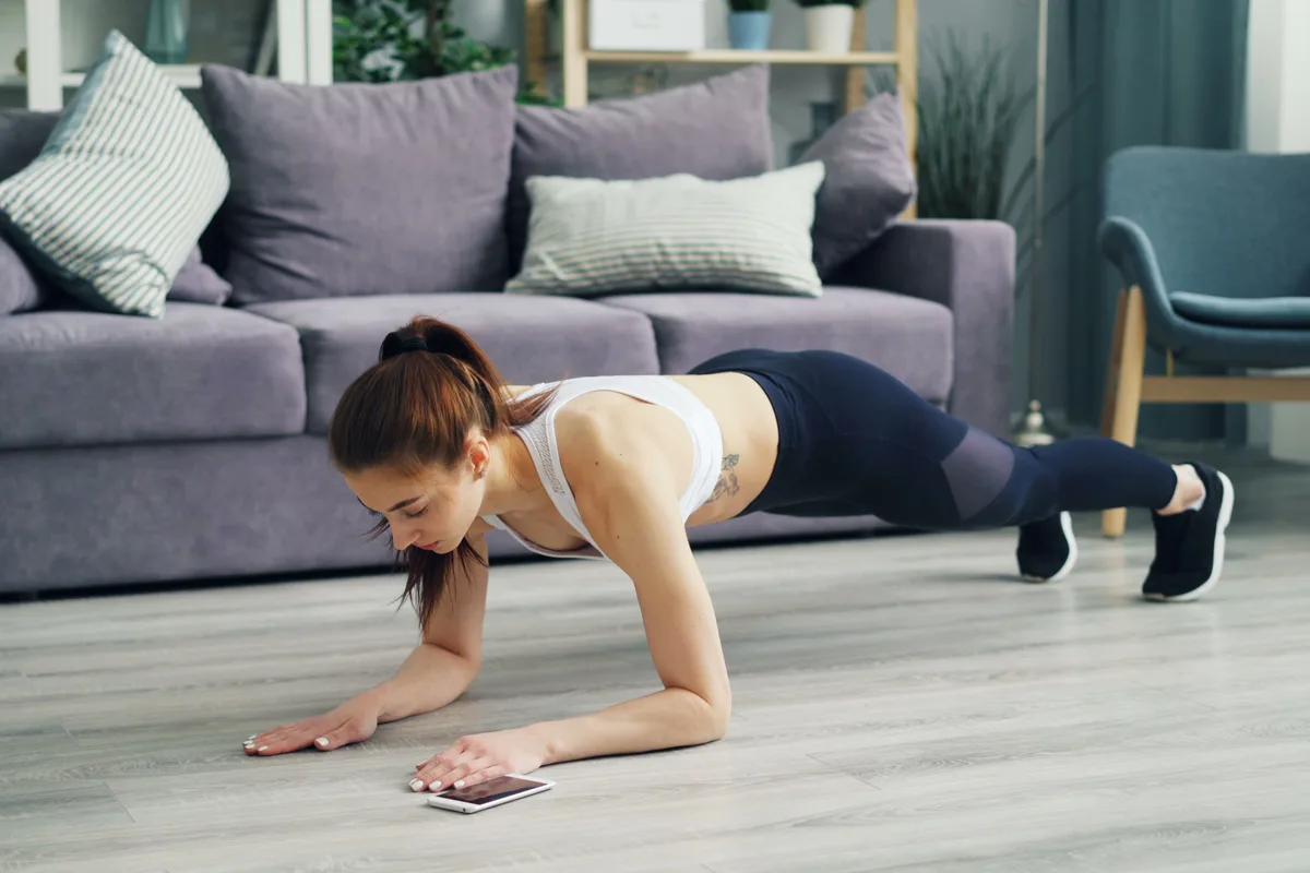 A woman holding a forearm plank on the wood floor of a bright living room, a phone on the floor by her hand for timing, with a grey sofa, striped cushions and a bookshelf behind her