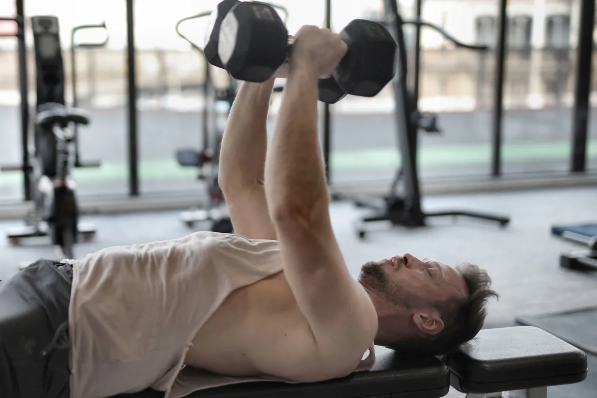 A man doing a dumbbell bench press on a bench in a bright gym, arms extended pressing the dumbbells above his chest