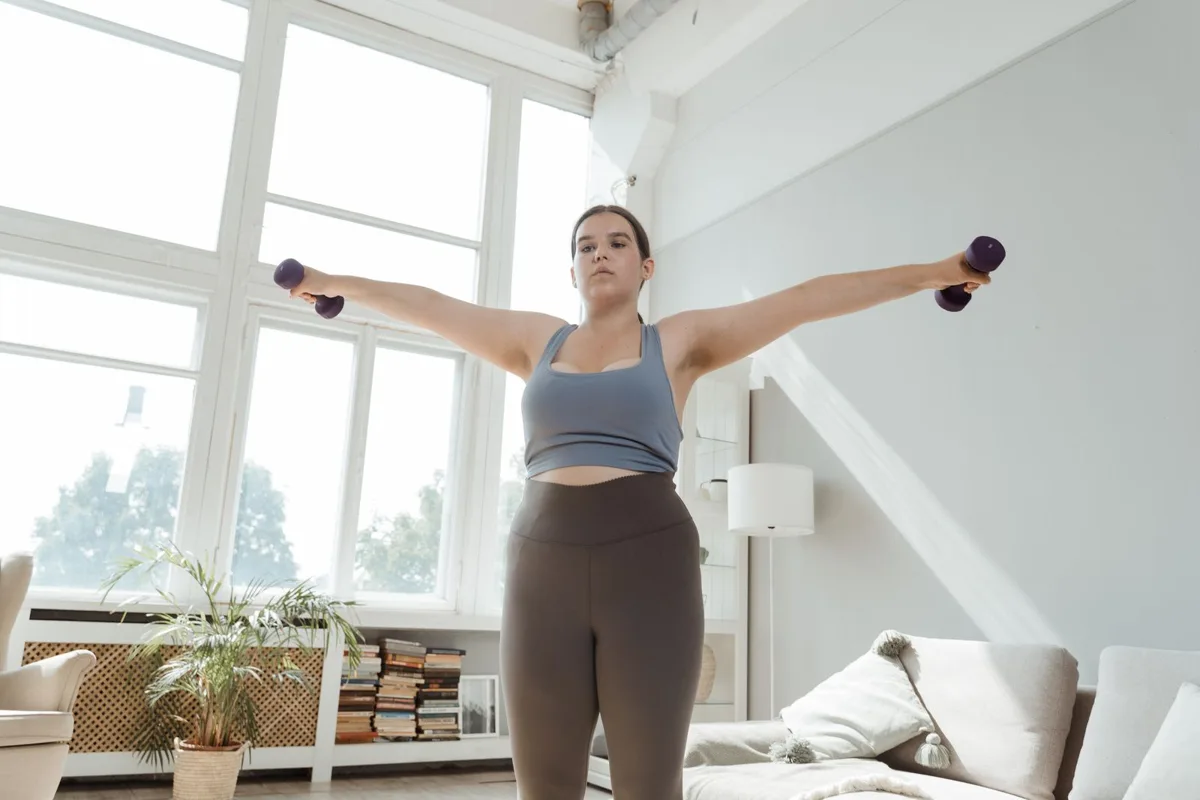 A woman doing a dumbbell lateral raise to shoulder height in a bright living room, arms out to the sides, with a beige sofa, bookshelves and plants behind her