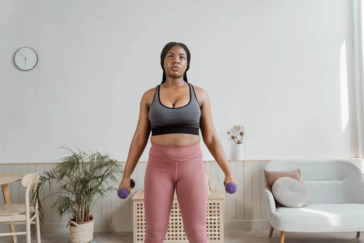 Dumbbell lateral raise starting position: standing in a bright living room holding a dumbbell in each hand at her sides