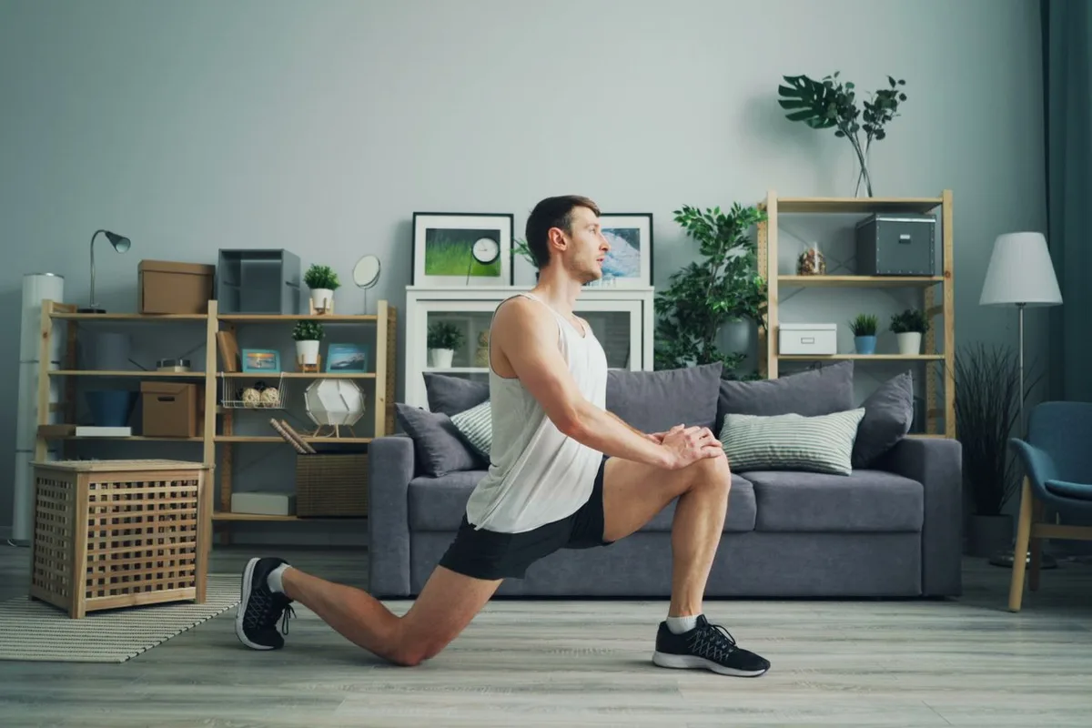 A man doing a lunge on the wood floor of a bright living room, one hand resting on his front knee, with a grey sofa, bookshelves and plants behind him