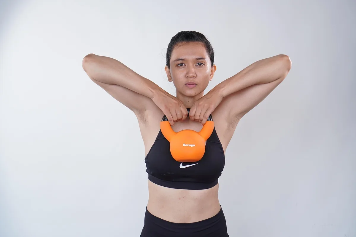 A woman holding an orange kettlebell with both hands up at her chest, elbows high, against a plain light background