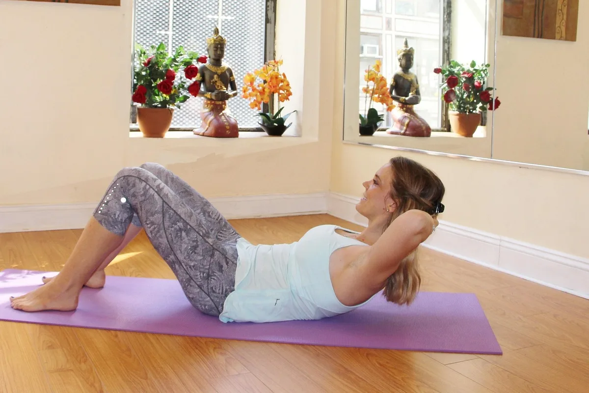 A woman doing a gentle core crunch on a lavender mat in a bright room with a large window, potted plants and ornaments on the windowsill, cream walls and a light wood floor