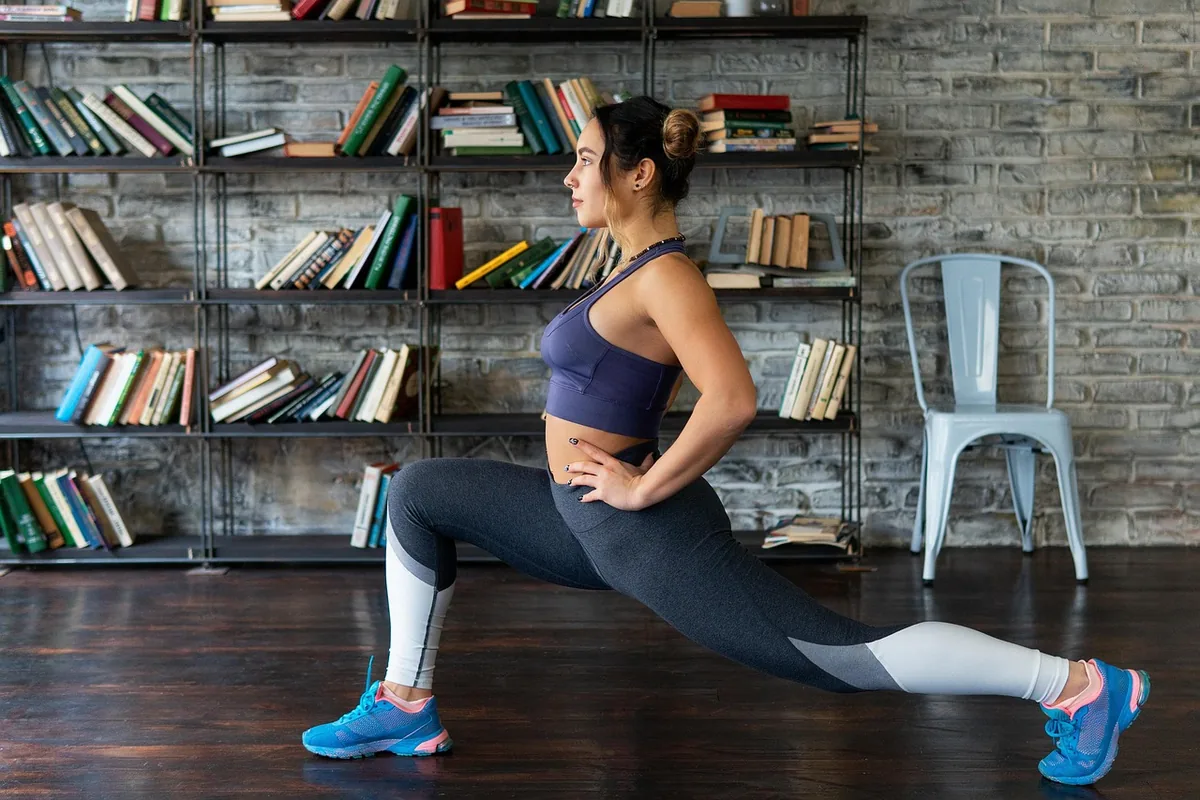 A woman doing a deep lunge in a home study in front of a full bookshelf, torso tall and hands on her hips, on a dark wood floor