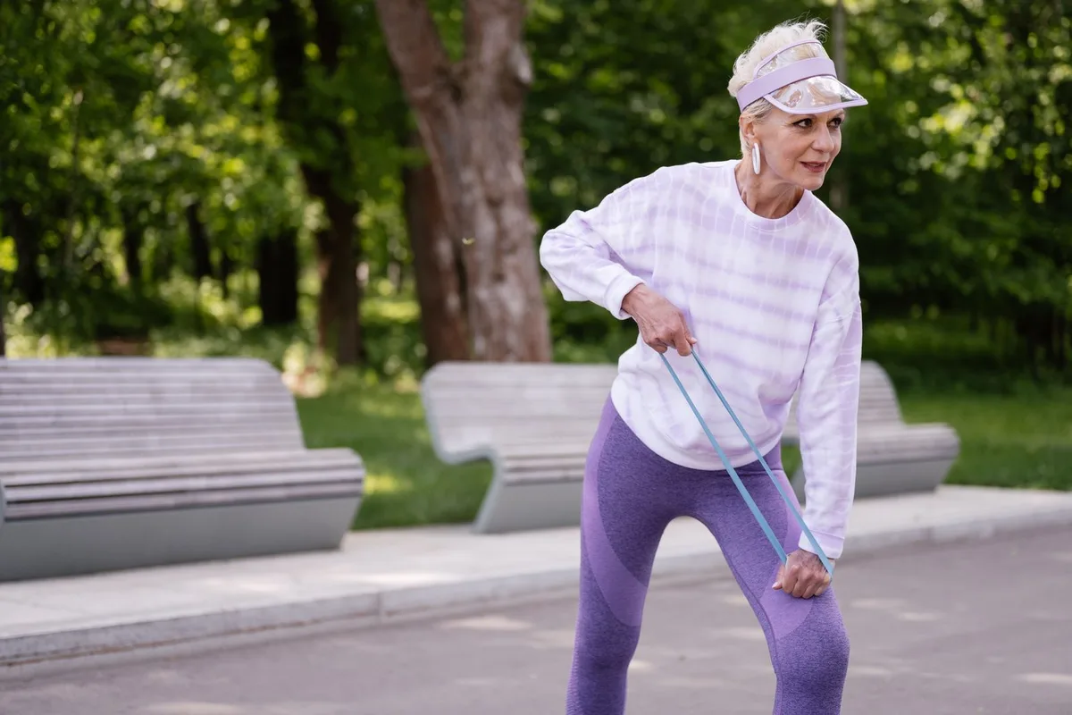 An older woman doing a one-arm resistance band row on a tree-lined park path, the band looped under her foot, with benches and green trees behind her